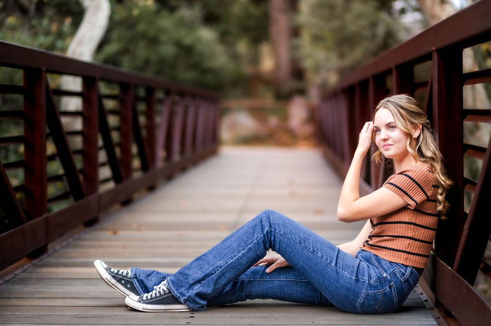 Young woman in jeans and striped top sitting on a wooden bridge outdoors.