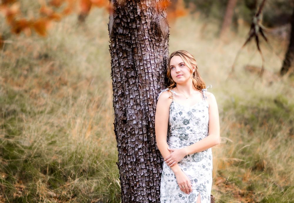 Young woman in floral dress leaning against a tree in a natural outdoor setting.