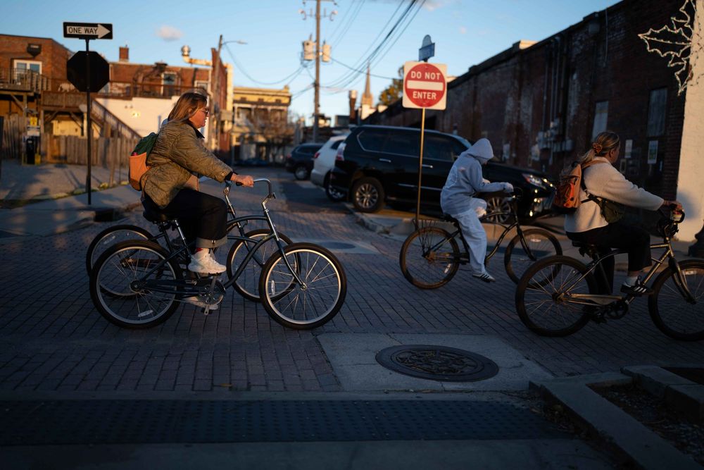 A sequence of cyclists riding through city streets at dusk with long shadows cast on the pavement.