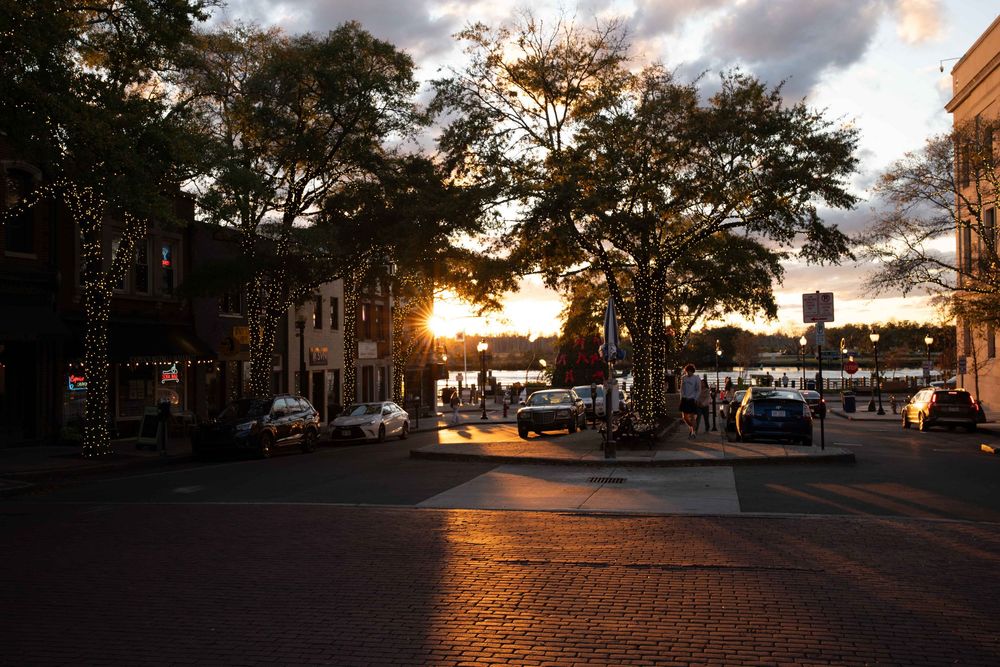 Sunset casts golden light across a brick paved street with historic buildings and trees silhouetted against the evening sky.