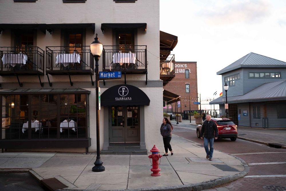 A street corner in a small downtown area with brick buildings, storefronts, and pedestrians walking on the sidewalk.