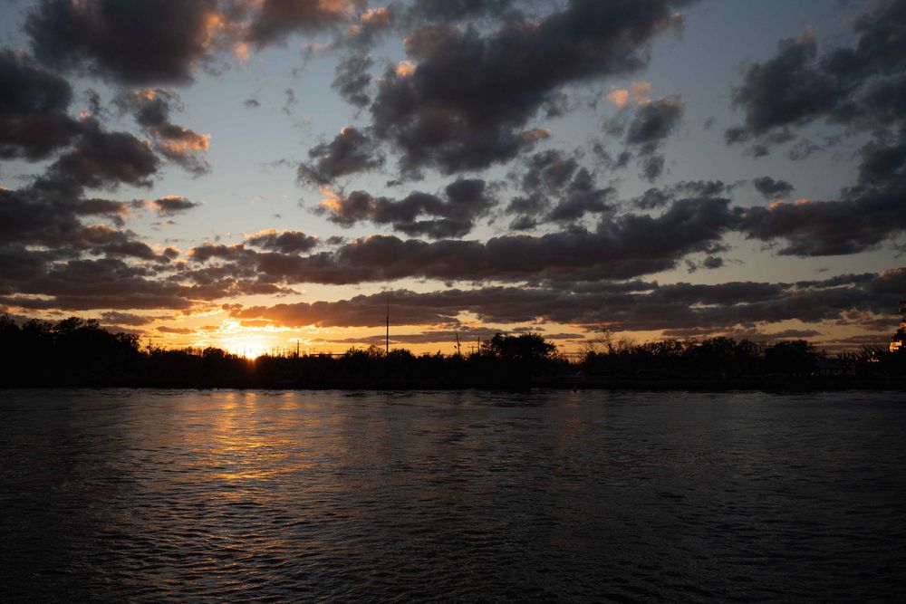 Dramatic dusk skyline with dark clouds and golden sunlight reflecting across a serene waterway at sunset.