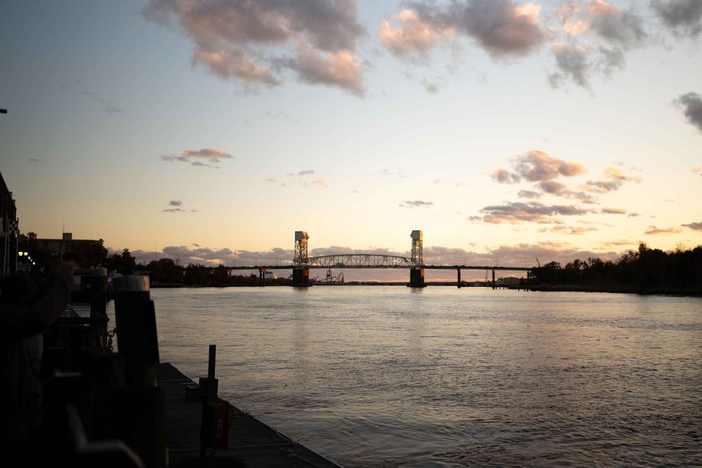 Panoramic sunset view over a river with silhouetted bridges and dramatic pink clouds reflecting on calm waters.