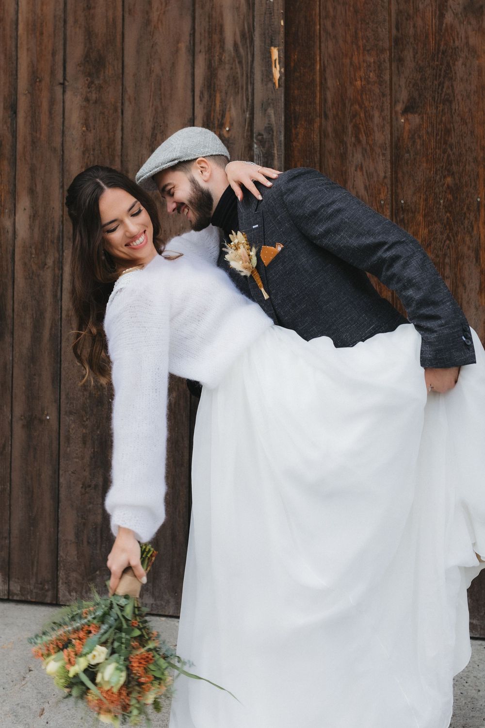 Playful wedding couple poses against rustic wooden barn wall with autumn bouquet.