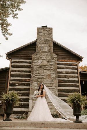 A bride in a flowing white gown stands in front of a rustic log cabin with a stone chimney.