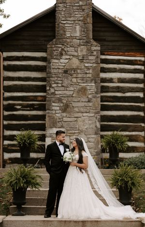 A wedding couple poses in front of a rustic log cabin with a grand stone chimney.