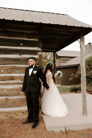 A wedding couple stands back-to-back outside a rustic wooden cabin with a covered porch.