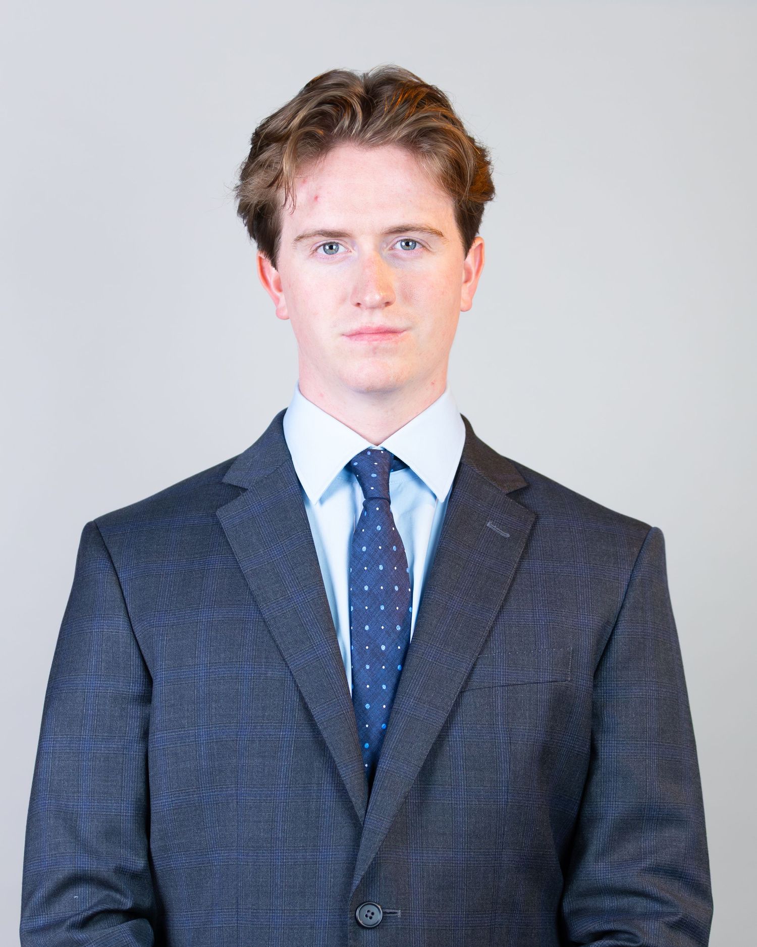 Professional headshot portrait of someone wearing a navy suit with polka dot tie against light background.