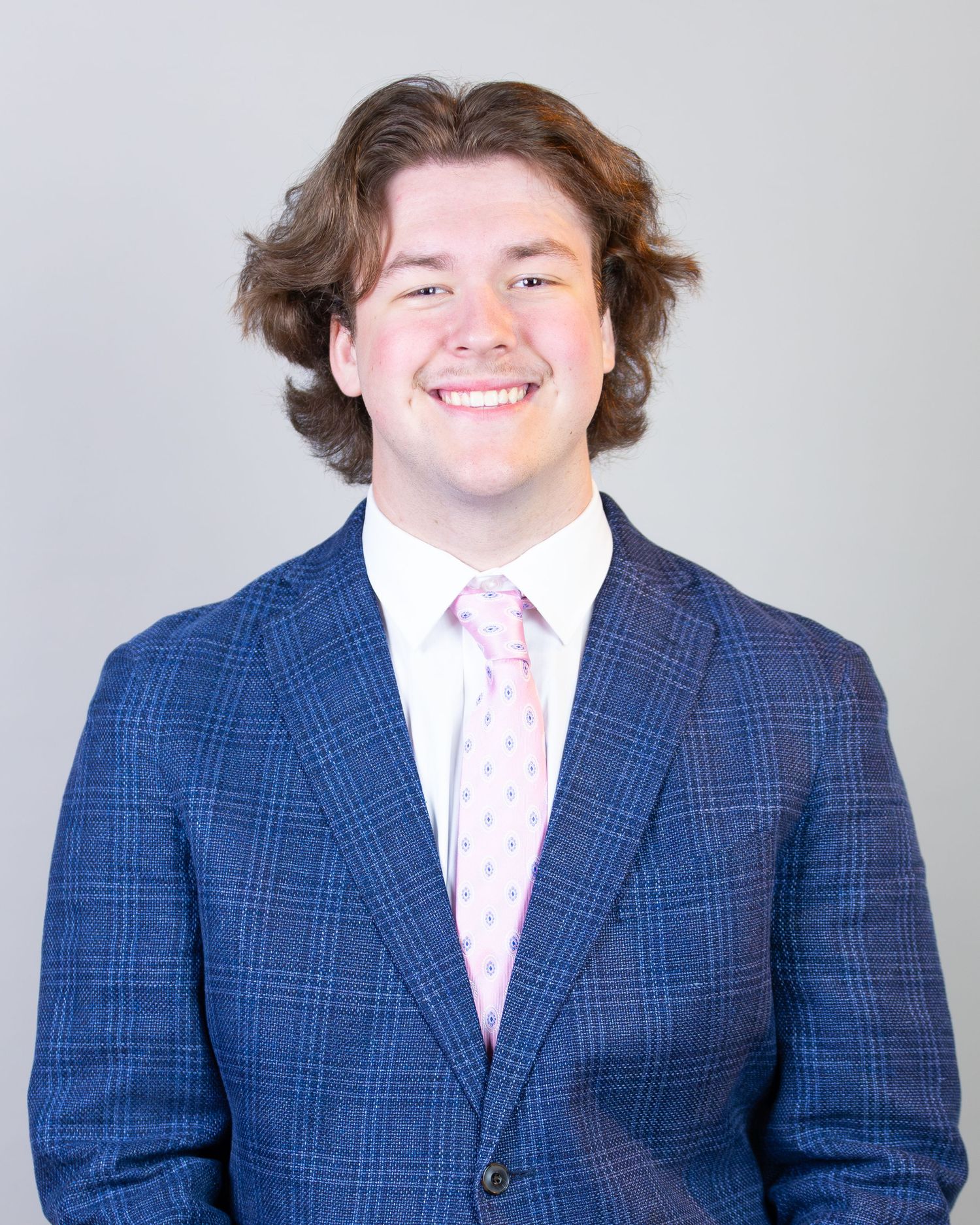 A person in a blue plaid suit jacket and pink striped dress shirt smiles warmly at the camera.