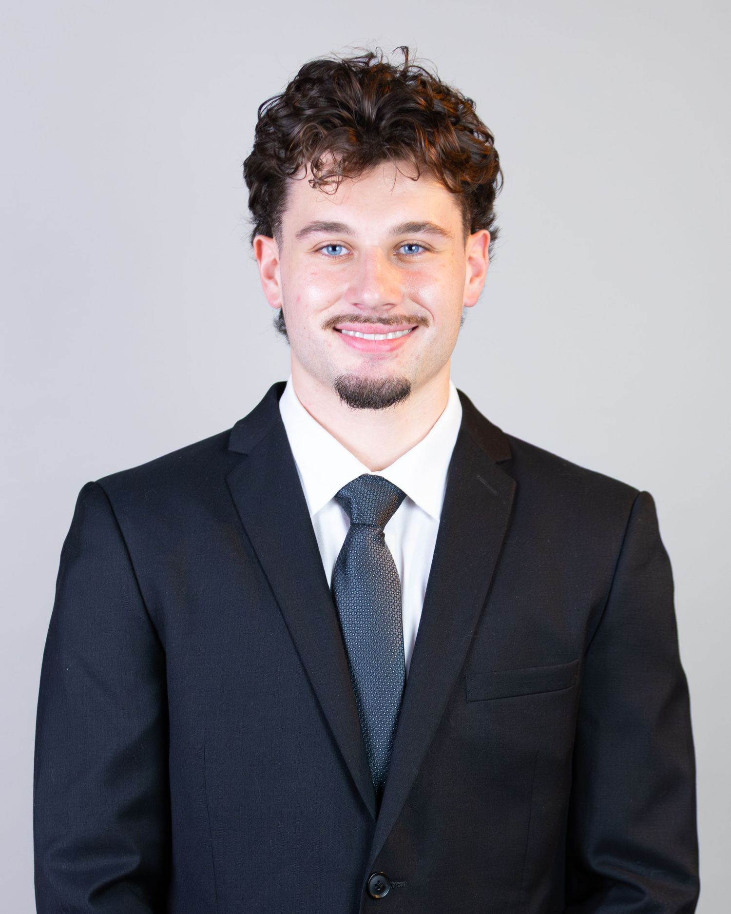 Professional headshot of someone in a black suit and tie against a light gray background.