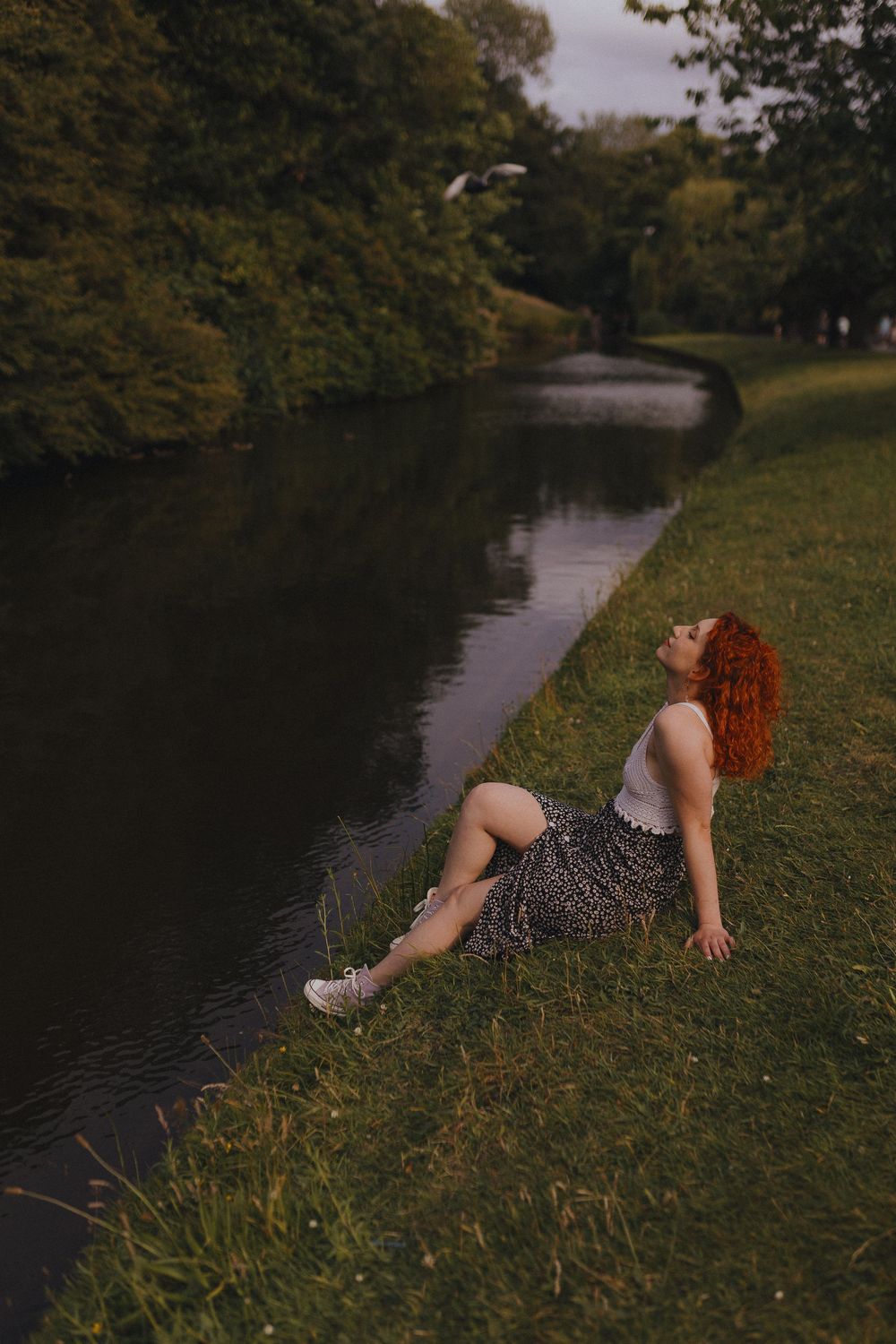A person in a polka dot dress lies on grassy riverbank at sunset creating a dreamy outdoor scene.