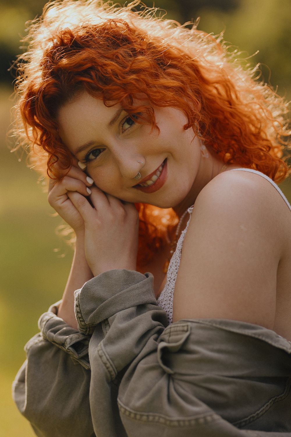 A woman with vibrant red curly hair poses playfully in a denim jacket against a warm golden outdoor background.