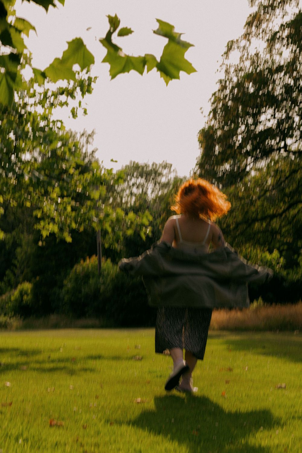 A figure in a dark dress twirls gracefully across a sunlit grassy field surrounded by trees.