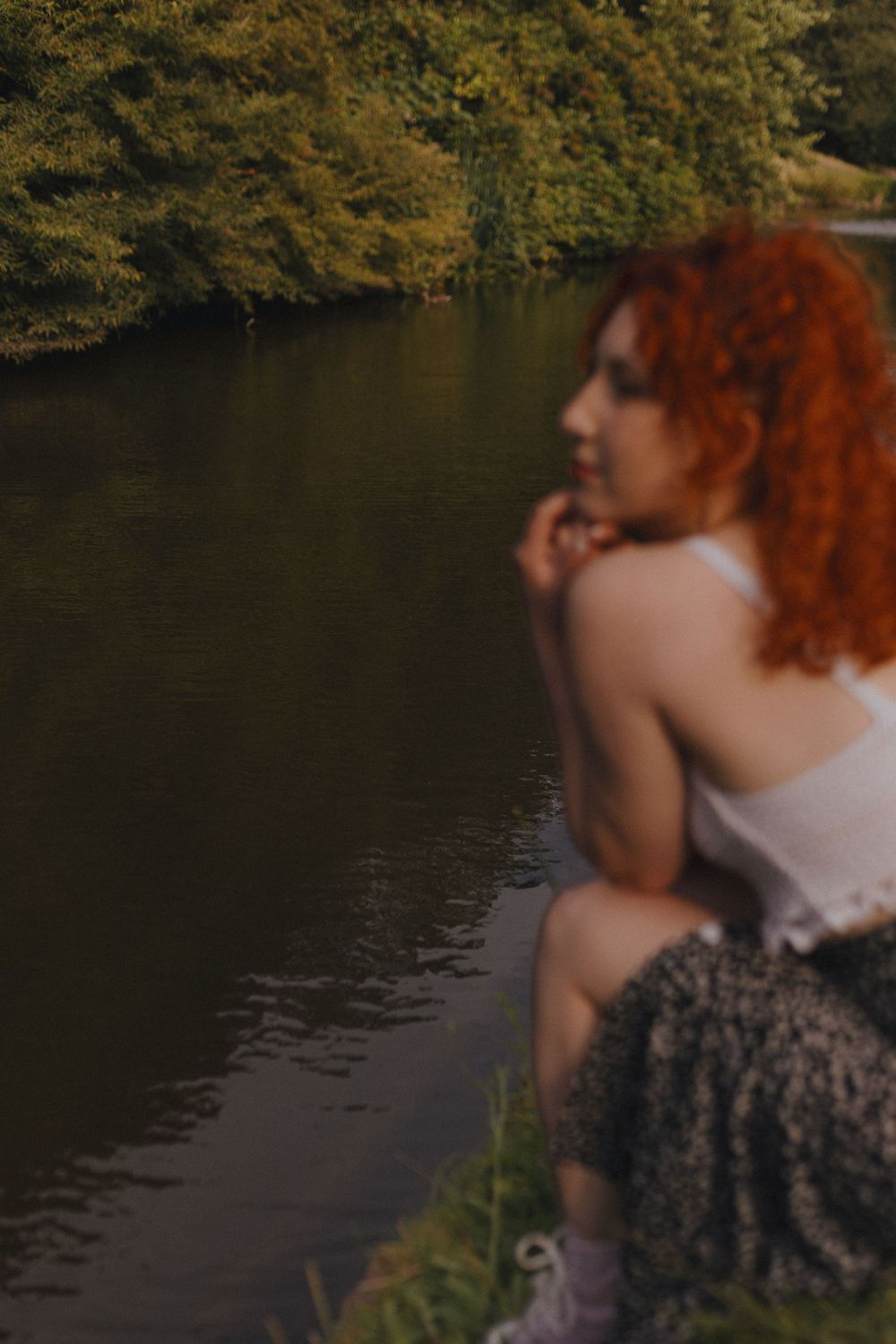 A person with curly red hair sits by a tranquil lake surrounded by autumn foliage.
