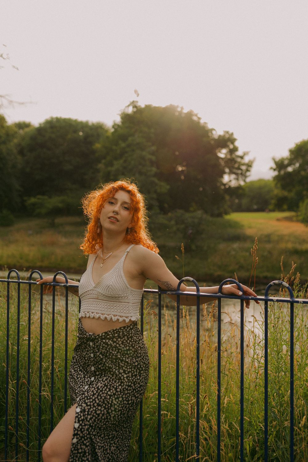 Person in a beige top and leopard print skirt poses against an iron fence at sunset in a grassy field.