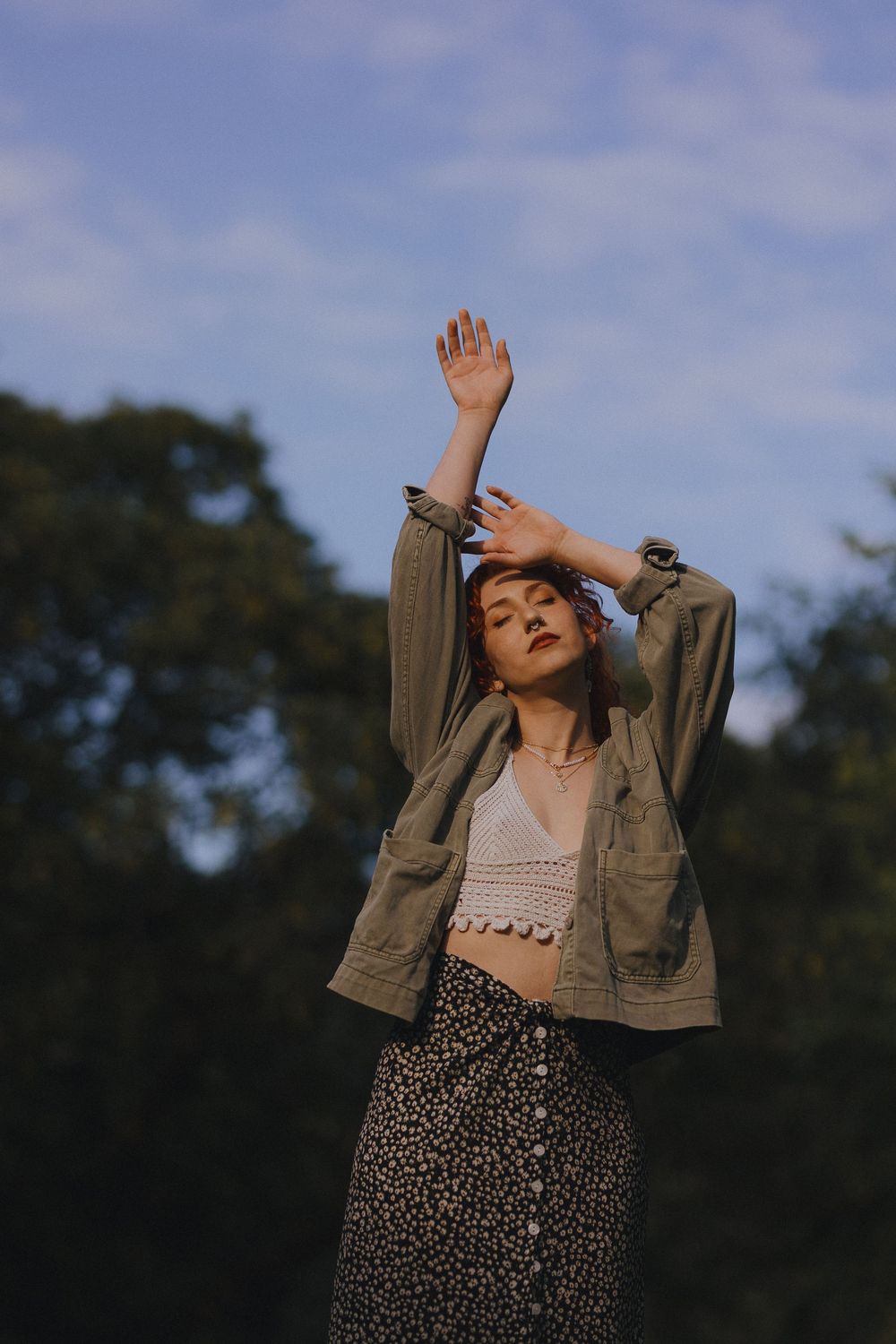 Person wearing an olive jacket and animal print skirt poses expressively against a blue sky backdrop.