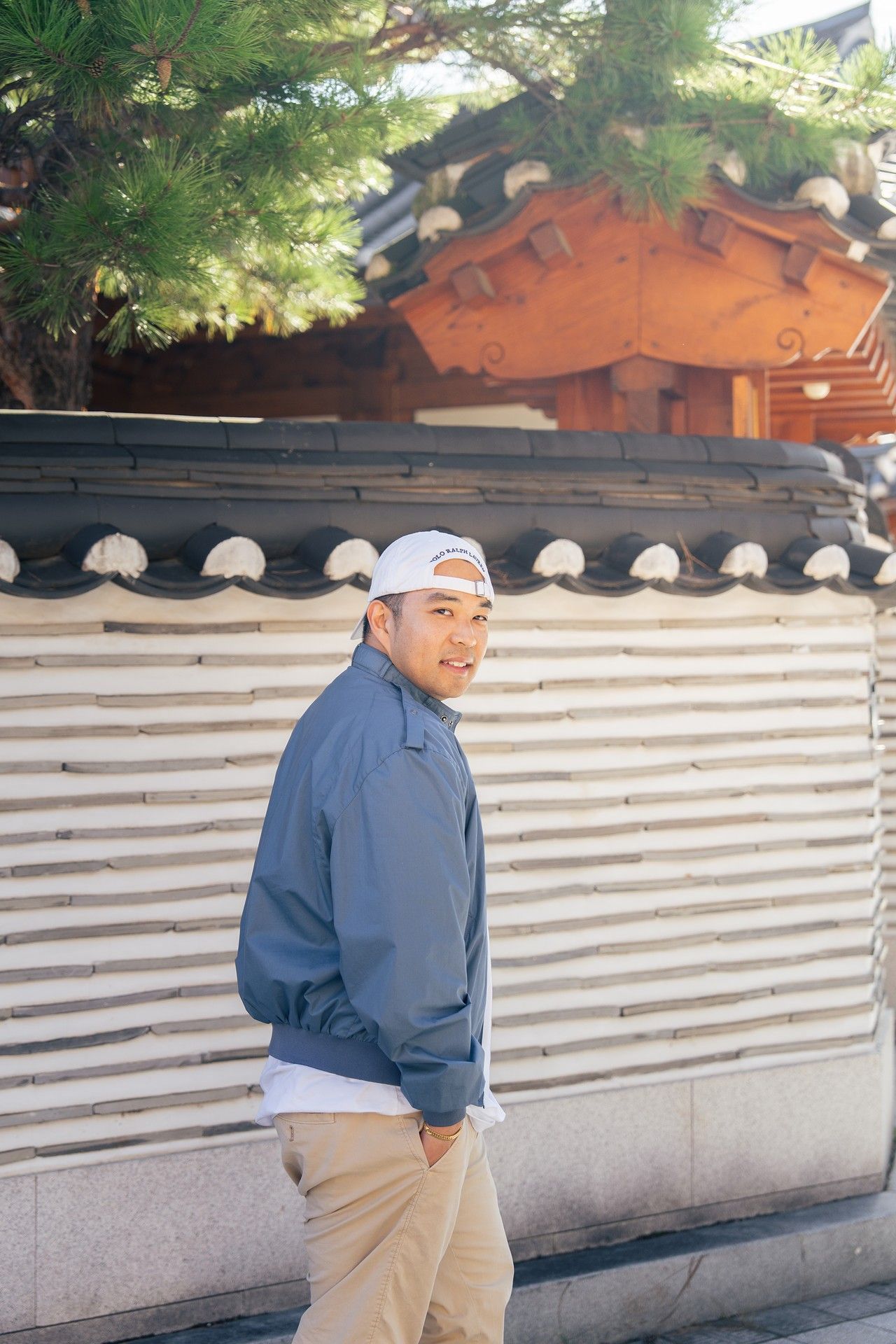 A person in a blue jacket and baseball cap walks past a traditional Korean stone wall with ornate roof tiles above.