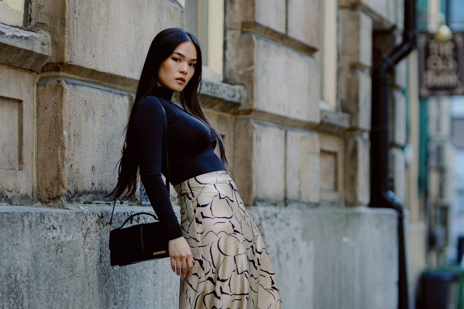 A woman in a black top and snake print skirt walks past a stone building facade on a city street.