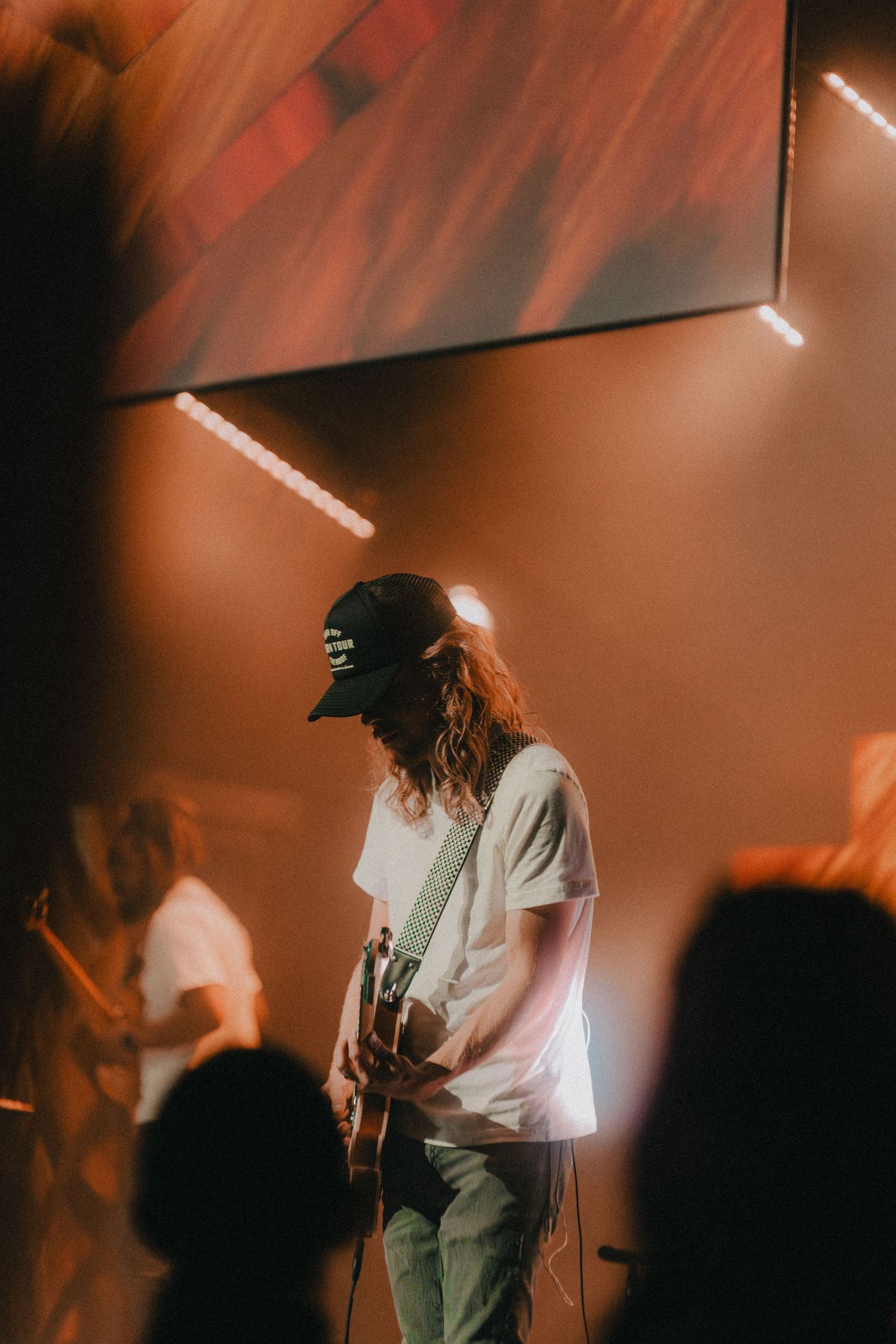 Concert performer in white t-shirt and baseball cap playing guitar on stage under dramatic red lighting.
