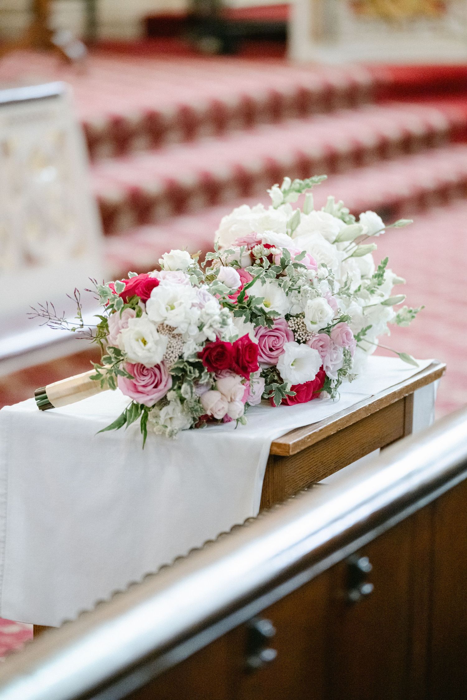 Pink and white floral arrangements with roses and greenery line a wooden church pew for a wedding ceremony.