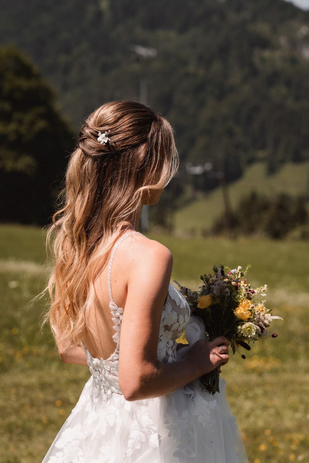 Bride in lace dress holding wildflower bouquet against mountain landscape.