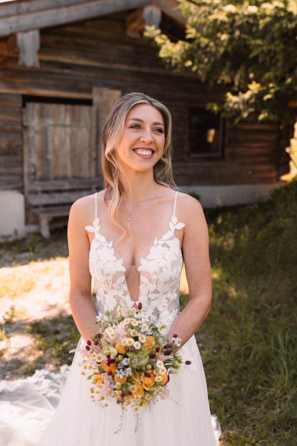 A bride in a lace wedding dress holds a vibrant yellow and orange wildflower bouquet while smiling outdoors.