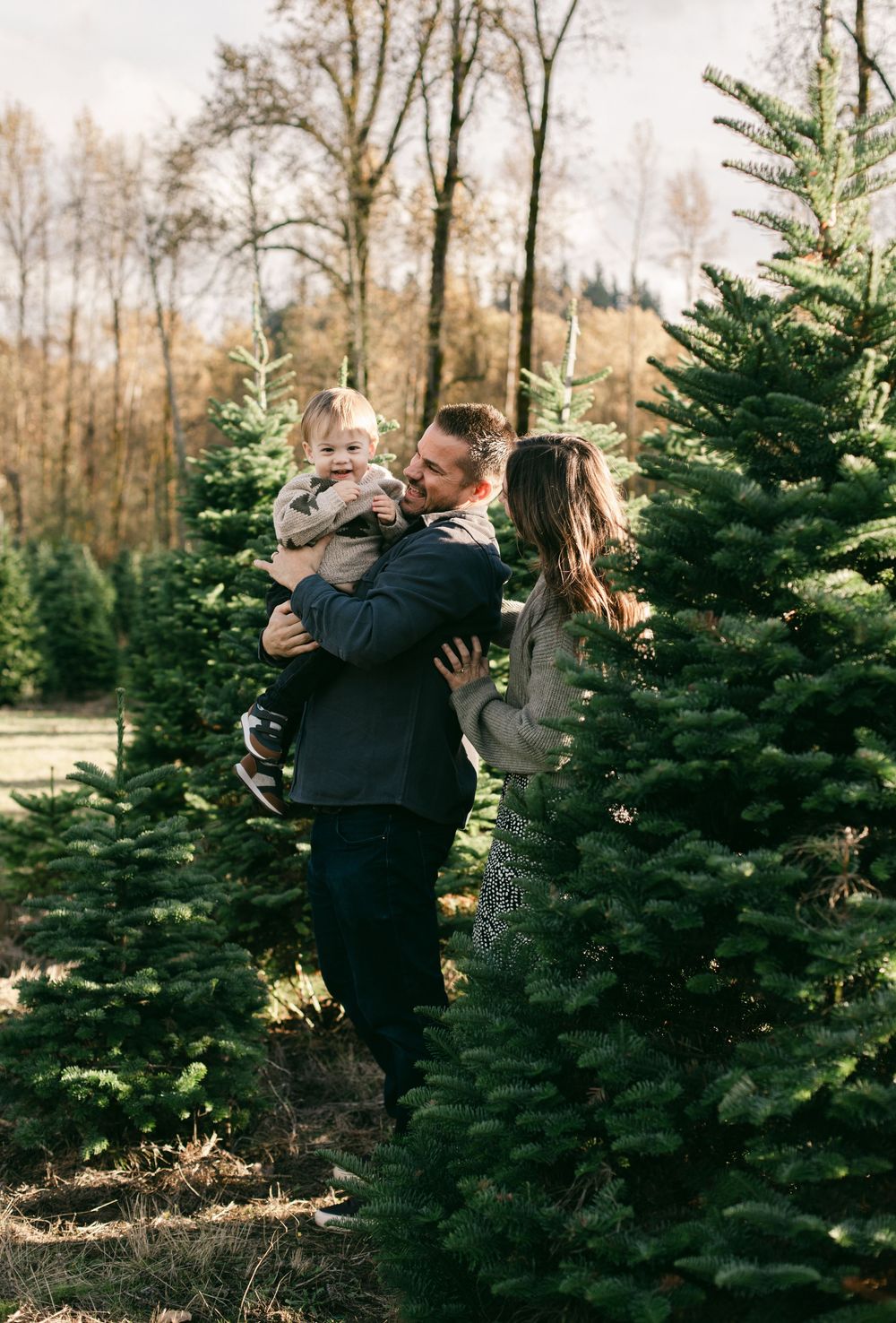 A family enjoys a winter outing at a Christmas tree farm surrounded by tall evergreen trees on a crisp autumn day.