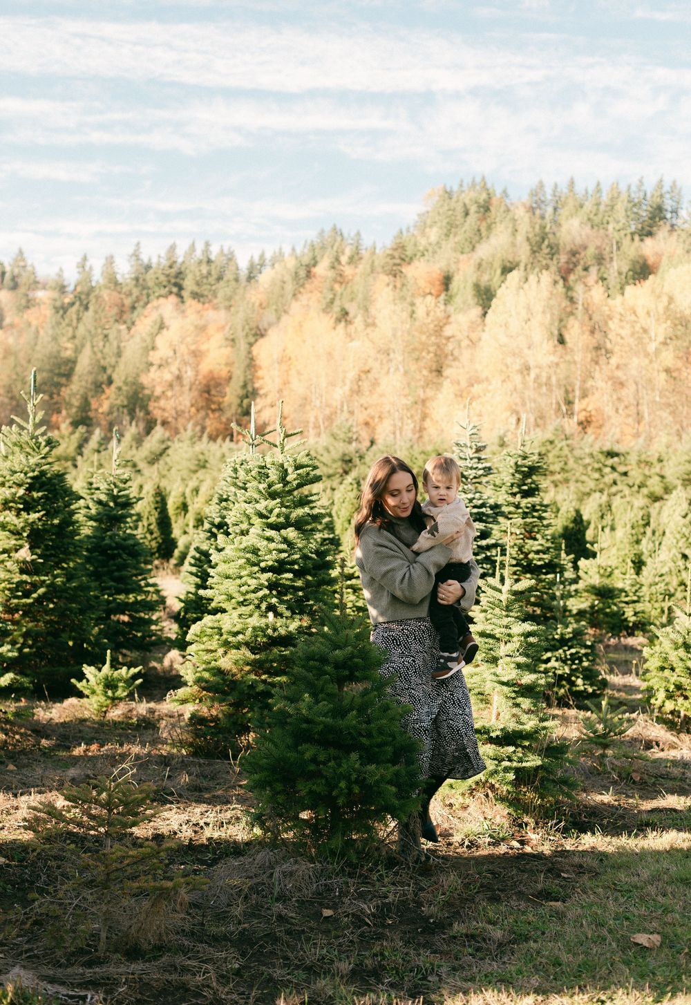Two people stand among evergreen trees at a Christmas tree farm on a sunny autumn day.