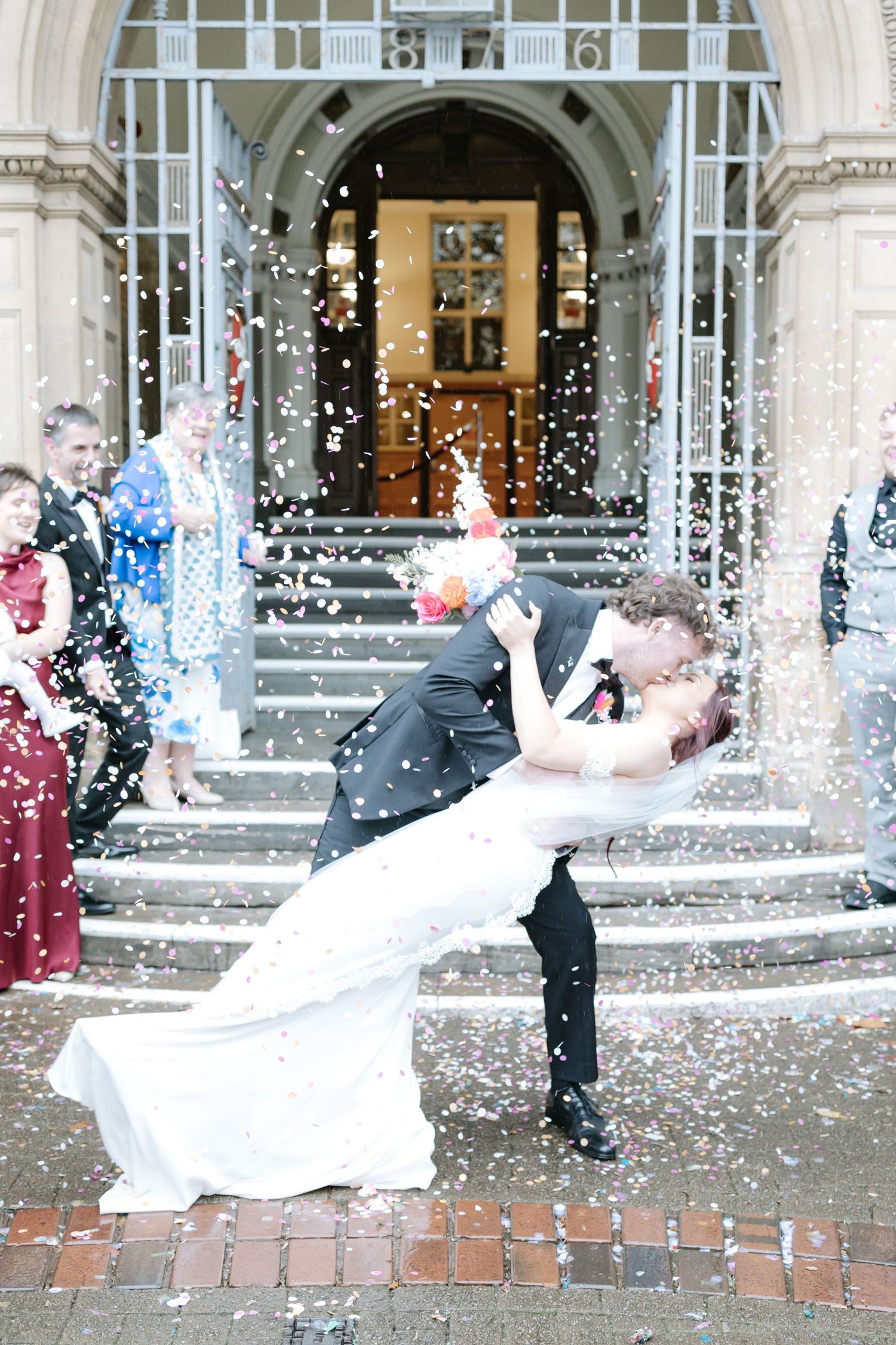 Newlyweds celebrate with confetti and joyful spins on brick steps of a historic building with elegant columns.