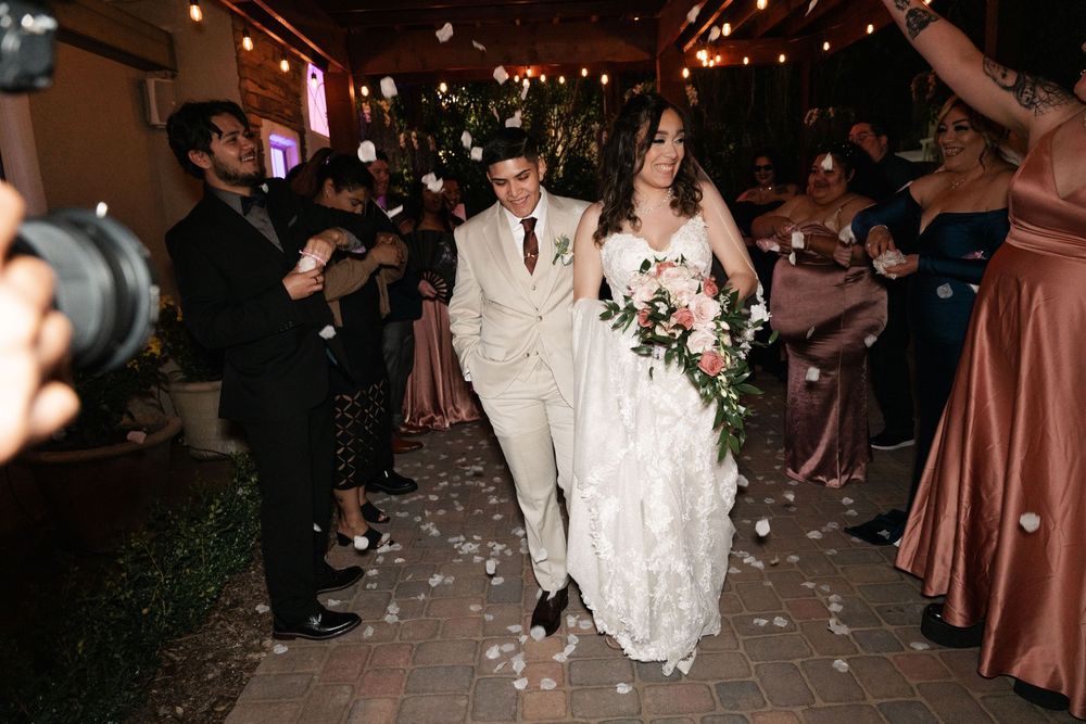 A newlywed couple walks down a candlelit path while guests throw flower petals during an evening wedding celebration.