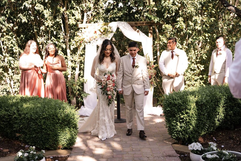 Wedding party members in beige suits and coral dresses stand together in a garden setting.