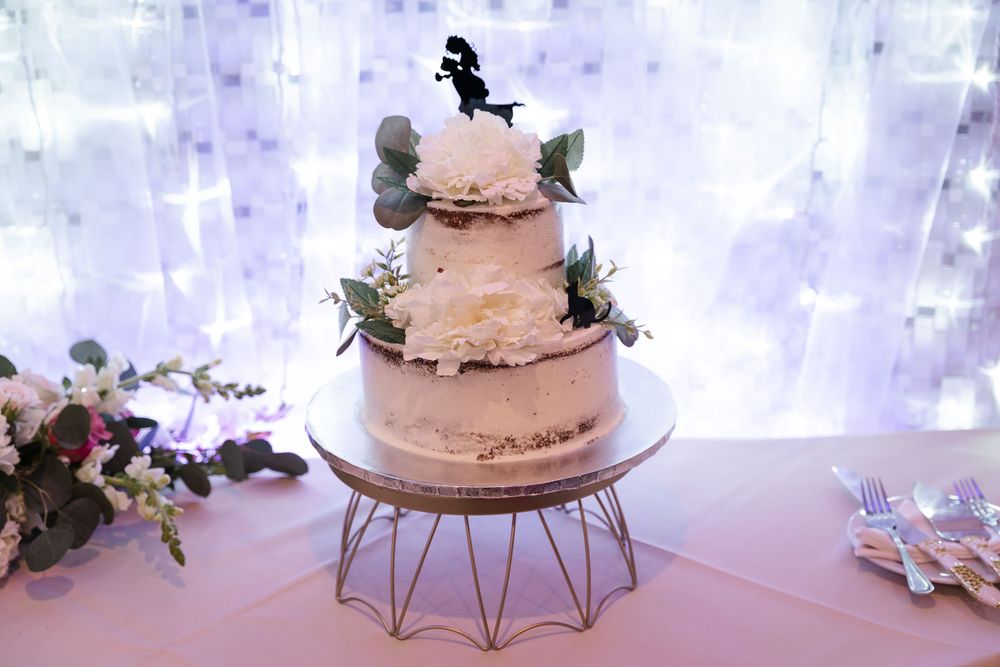 Three-tiered wedding cake with silhouette topper displayed on modern wire stand with twinkle light backdrop.