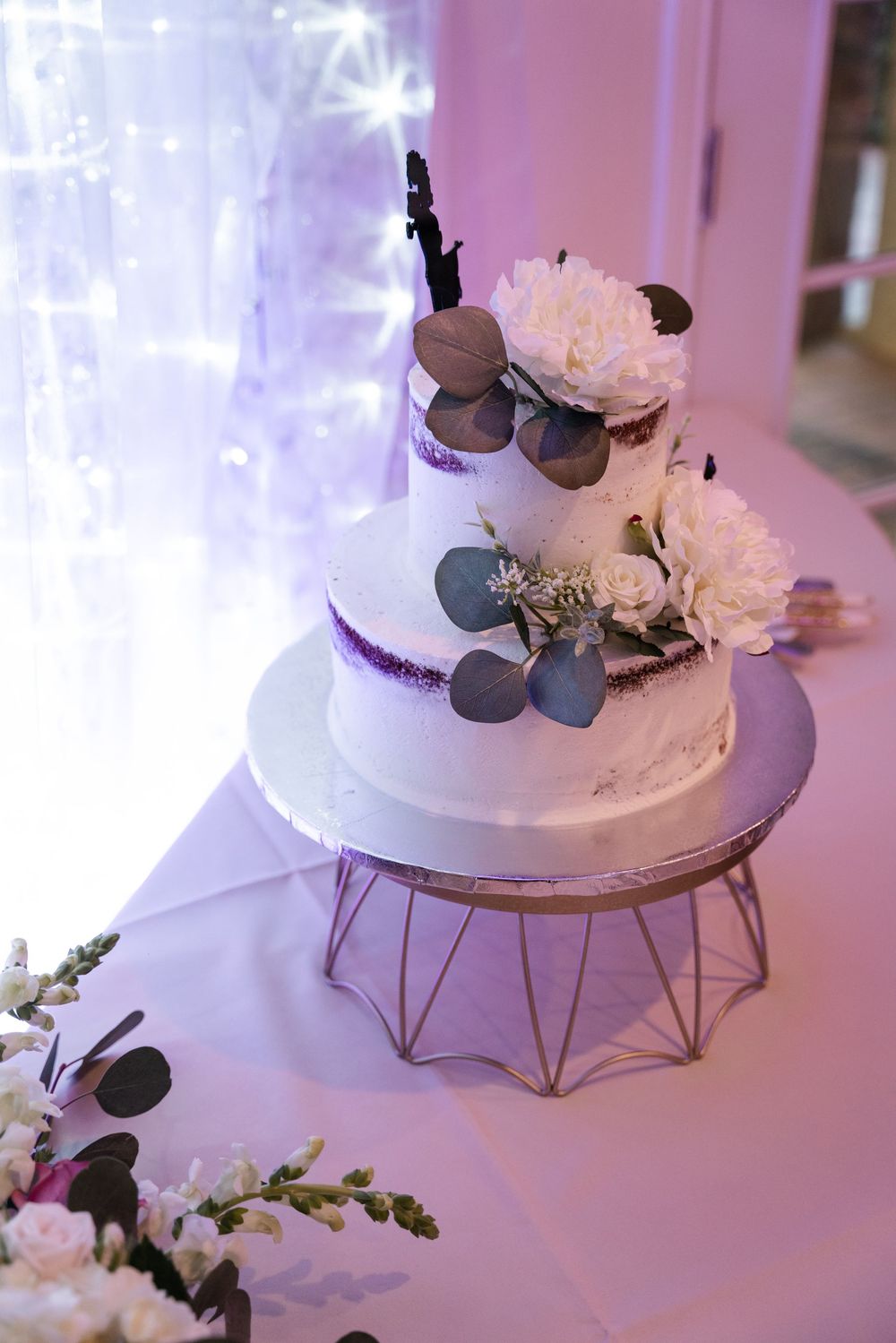 Three-tiered white wedding cake decorated with fresh flowers and greenery on a geometric metal display stand.