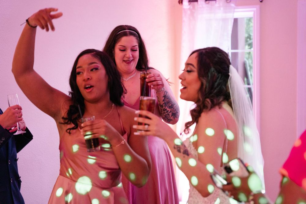 Group of friends enjoying drinks and conversation at a pink-lit party with glowing string lights in background.