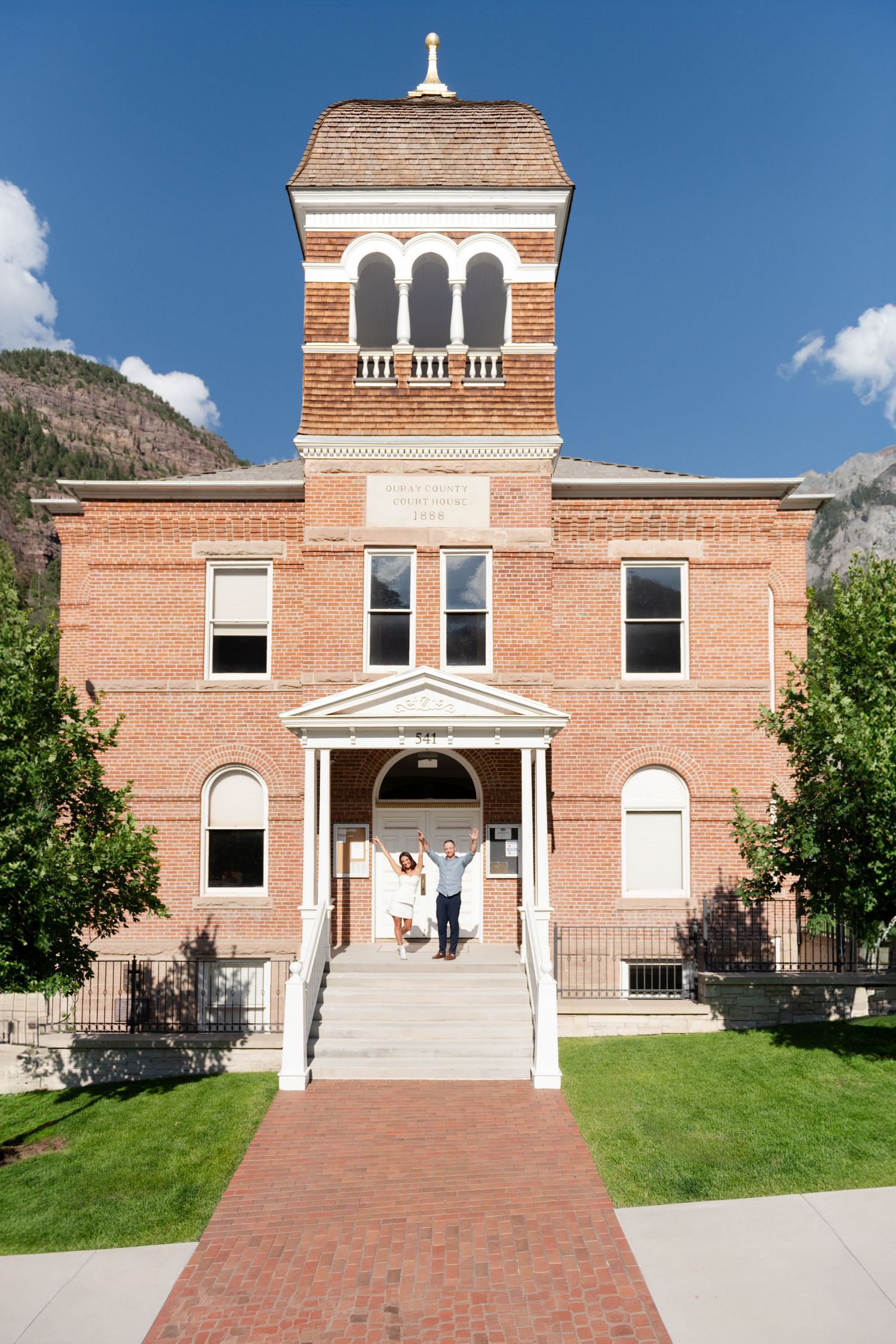 An eloping couple celebrate on the steps of the Ouray county courthouse. She is in a short white dress and he is soft blue shirt and pants.