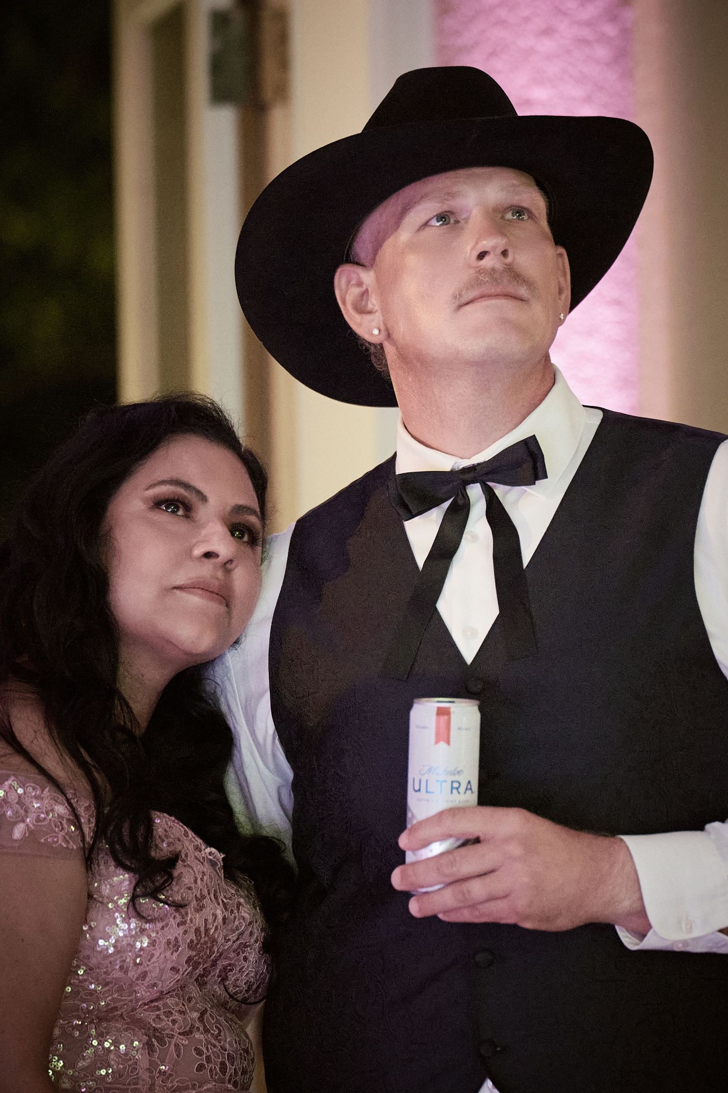 A couple in formal attire shares an intimate moment at an event, with the person wearing a black cowboy hat and tie.