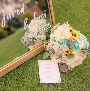 Wedding details with invitation and flowers arranged on grass with golden frame.