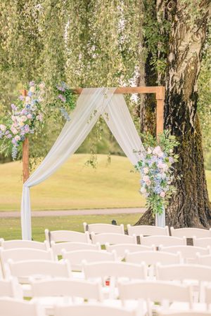 Outdoor wedding ceremony setup with white chairs and floral arch under weeping willow trees.