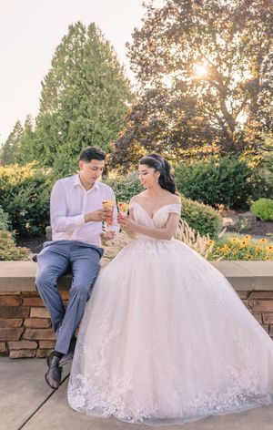 A couple in wedding attire shares a romantic moment on a stone wall at sunset.