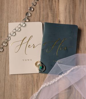 His and her vow books with gold lettering displayed with wedding accessories on wooden background.