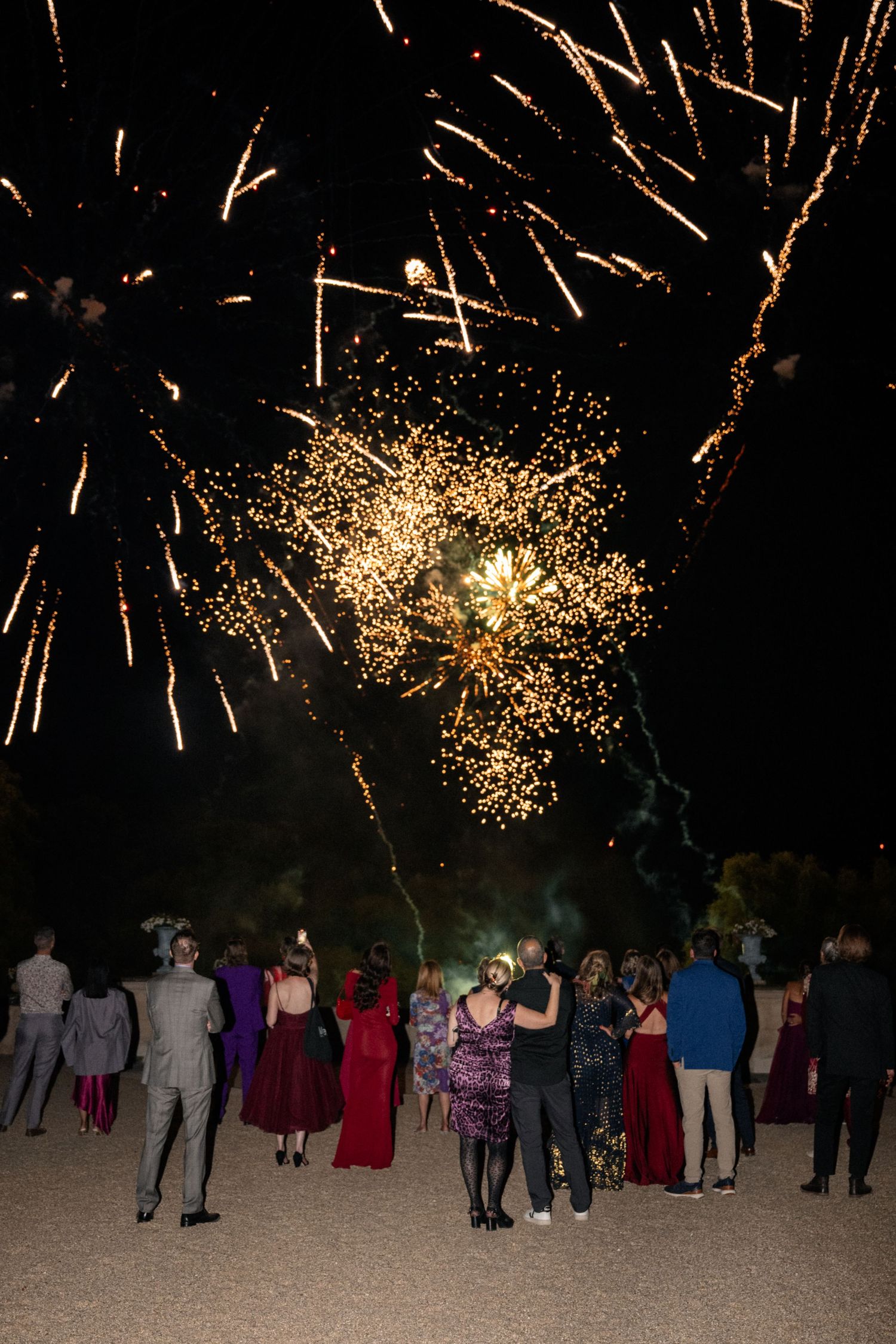A crowd of people watch golden fireworks explode in the dark night sky during an outdoor celebration.