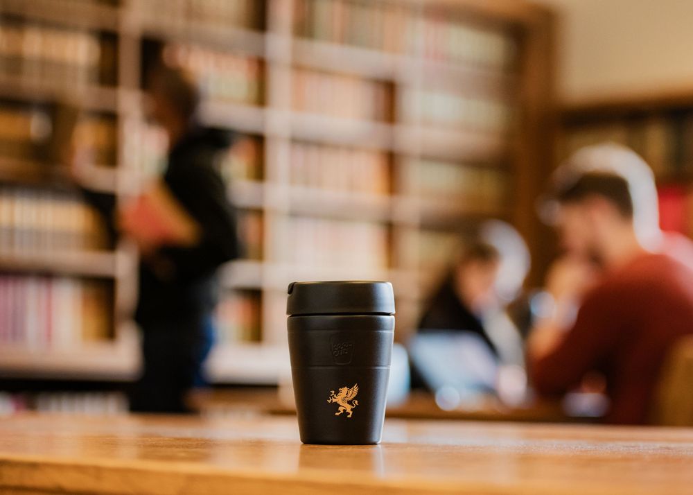 Starbucks coffee cup sits on wooden table in blurred cafe interior with bookshelves in background.