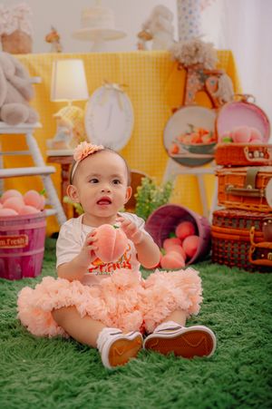 Baby wearing peach tutu dress sitting on green carpet with decorative props.