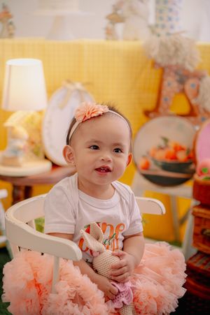 Baby in peach tutu dress sitting in white chair against yellow backdrop.