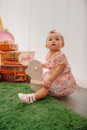 Baby in pink floral outfit sitting on green carpet near decorative boxes.