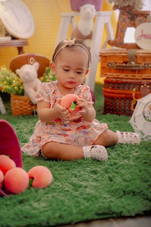 Baby in floral dress exploring toys and props on green carpet setting.
