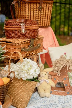 Picnic setup with wicker baskets, white flowers, and straw bags arranged on a lace tablecloth in an outdoor setting.