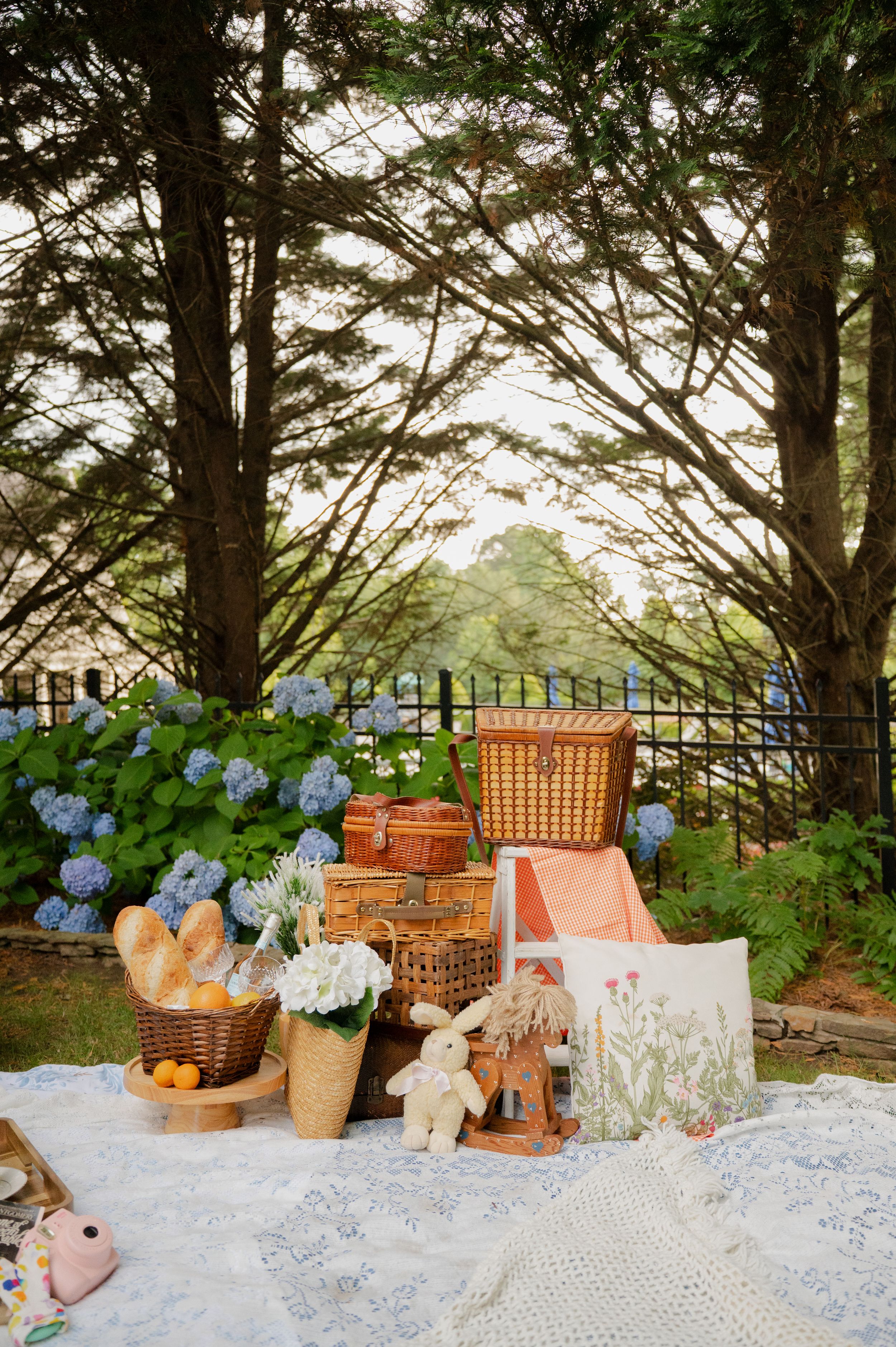 Garden picnic scene with hydrangeas, wicker baskets, and white linens under pine trees.