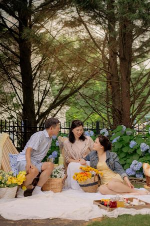 Summer picnic setup with fresh fruit, flowers, and wicker baskets on a white blanket under trees.