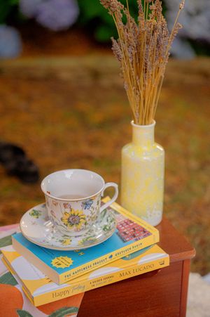 A vintage teacup and stack of yellow books with dried flowers in a vase on wooden surface.
